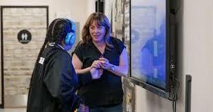 Teacher Alyson Leos smiles warmly while holding hands with a student wearing headphones in a classroom setting, standing beside an interactive display screen.
