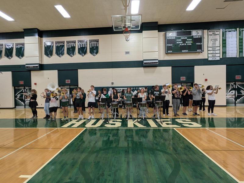 School band playing instruments on the gym floor, with a focus on the word 'Huskies.'