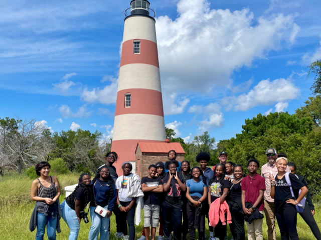 Group of Marine Science student posing in front of the Sapelo Island lighthouse under a blue sky.