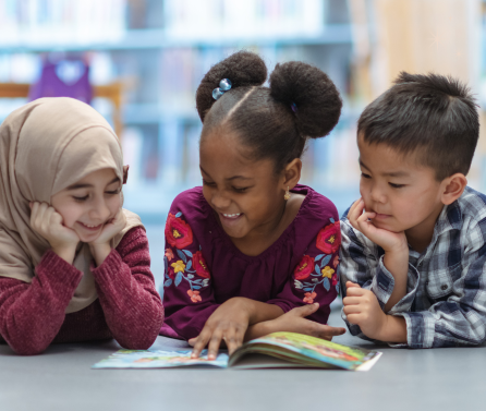 Three young children lying on the floor smiling and reading a book together in a library setting