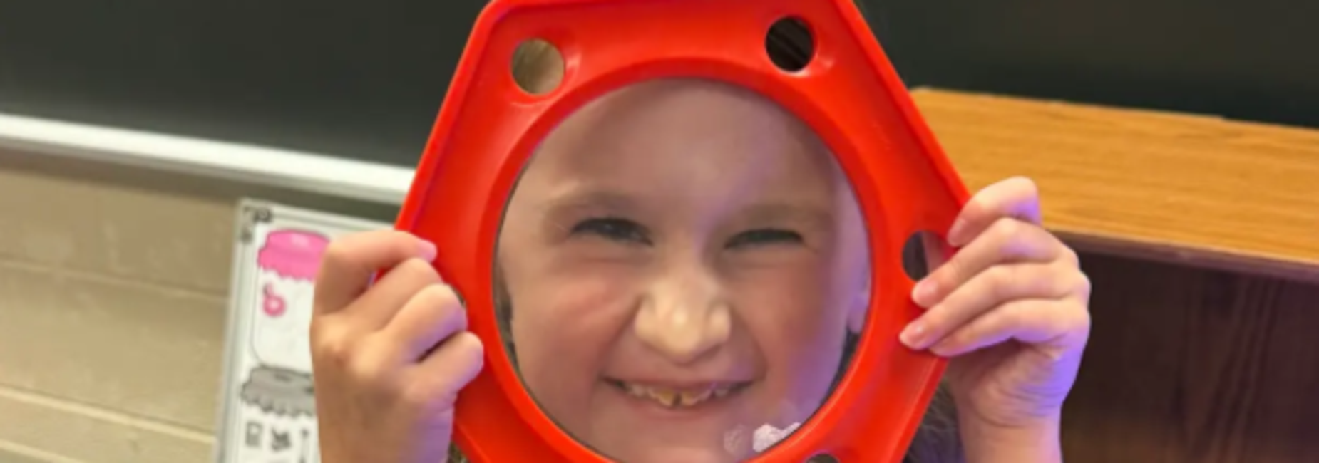 Child holding a red magnifying glass, smiling and showing teeth.