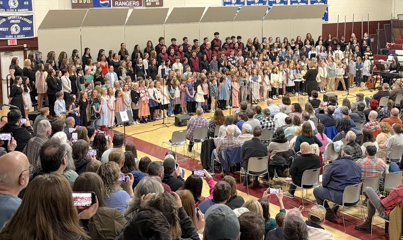 A large choir performance with children and adults on stage in a gymnasium.