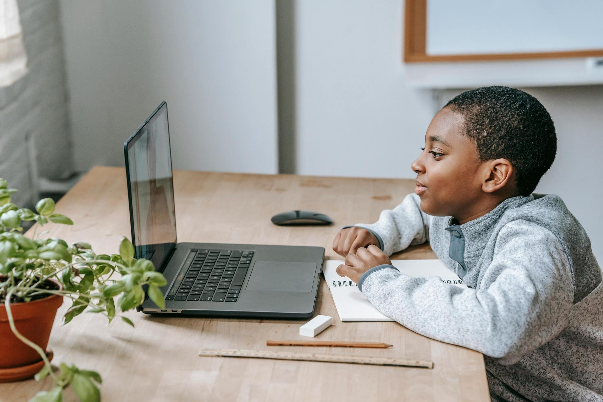 A boy sits at a table, engaged in an online meeting on his laptop.