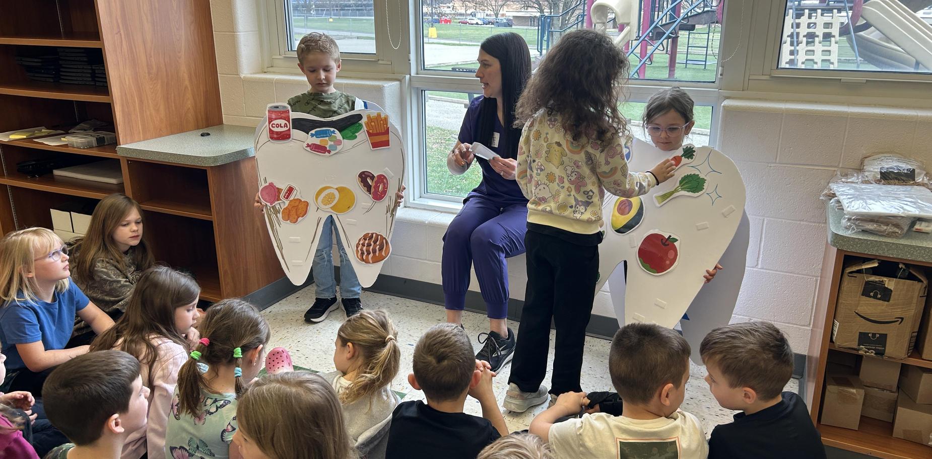 Children seated in a classroom watching a teacher demonstrate healthy foods on large models.
