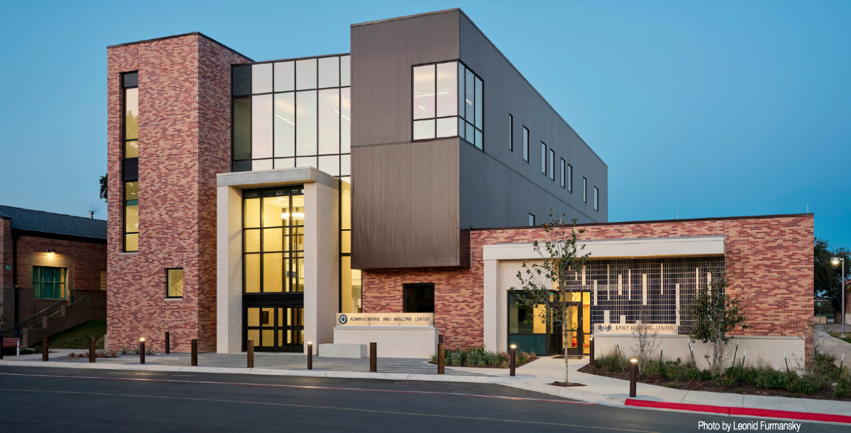 The Texas School for the Deaf (TSD) Administrative and Welcome Center, a modern three-story building with a mix of red brick and dark metal panels, illuminated warmly at dusk. Large windows provide a view of the building’s interior, showcasing a welcoming and contemporary design. The adjacent Early Learning Center is visible, surrounded by landscaped greenery, creating an inviting and professional campus atmosphere.