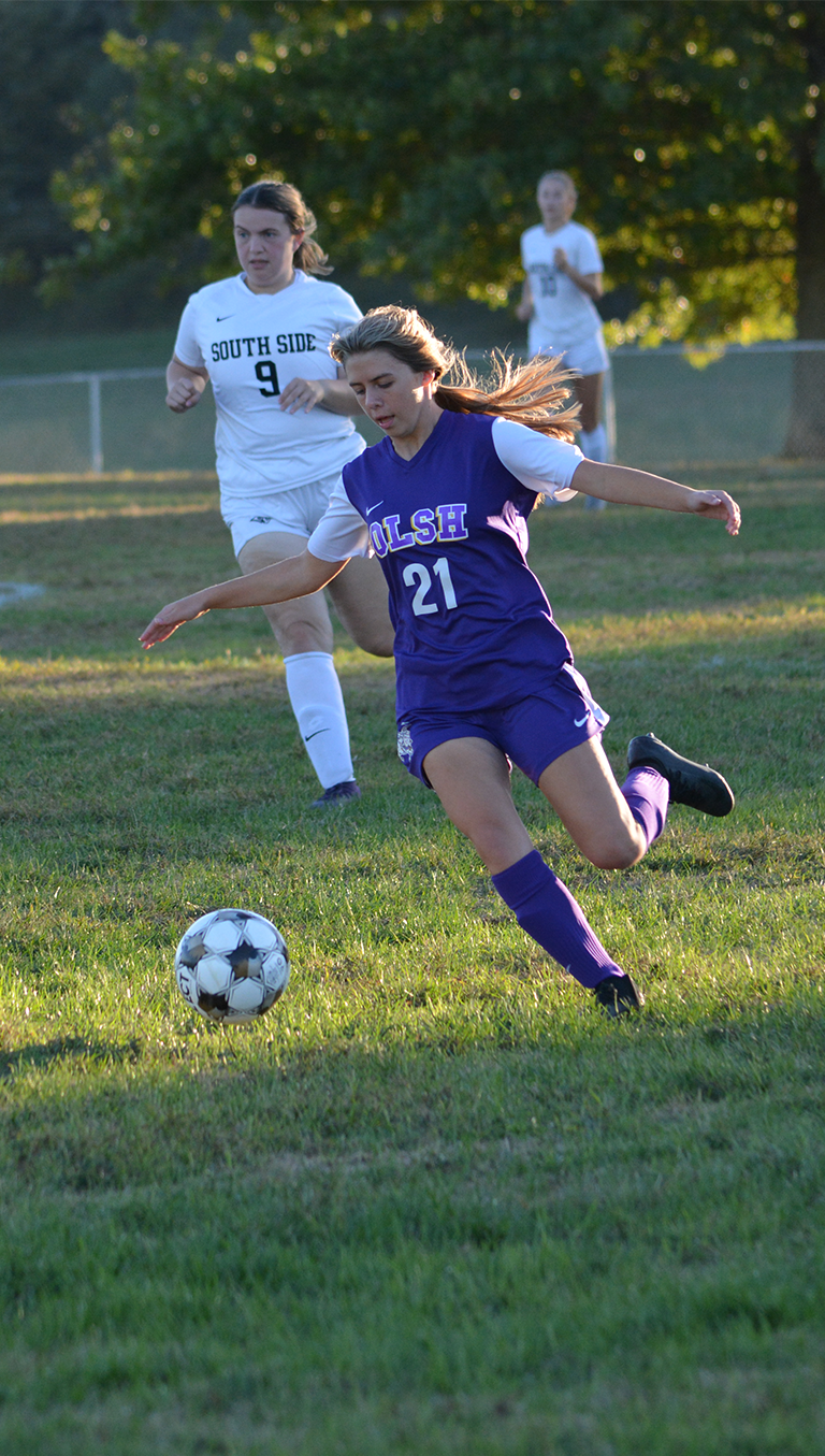 girls soccer player in purple uniform prepares to kick ball as opponents in white uniforms approach