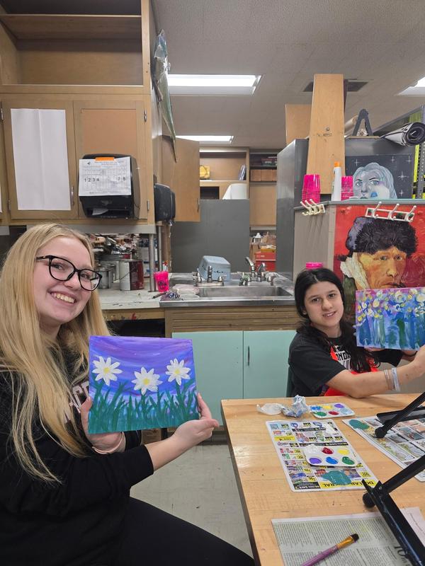 Two young women proudly displaying their flower paintings in an art studio.