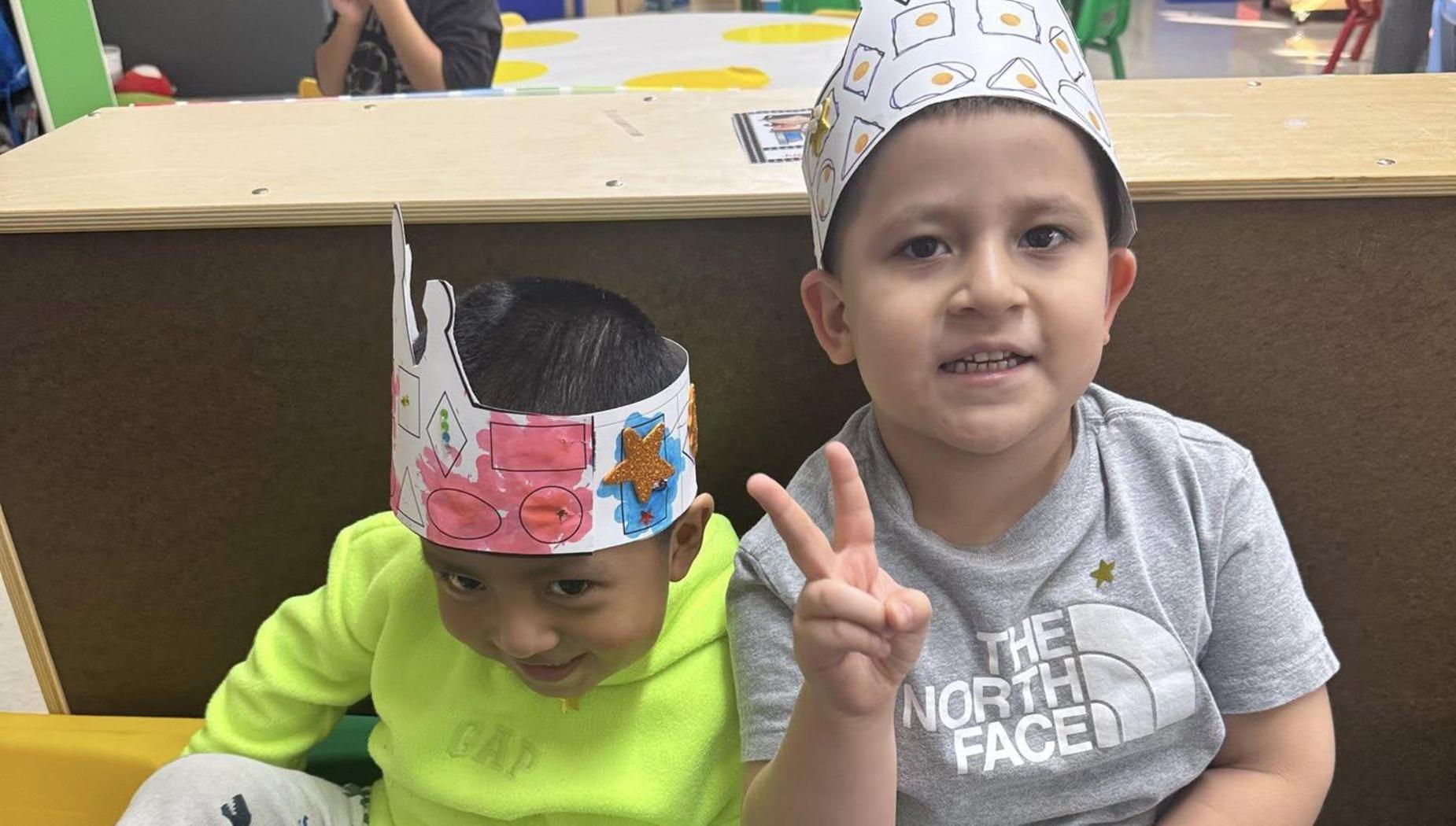 Two boys sitting together wearing paper crowns and giving peace signs, with playful expressions.