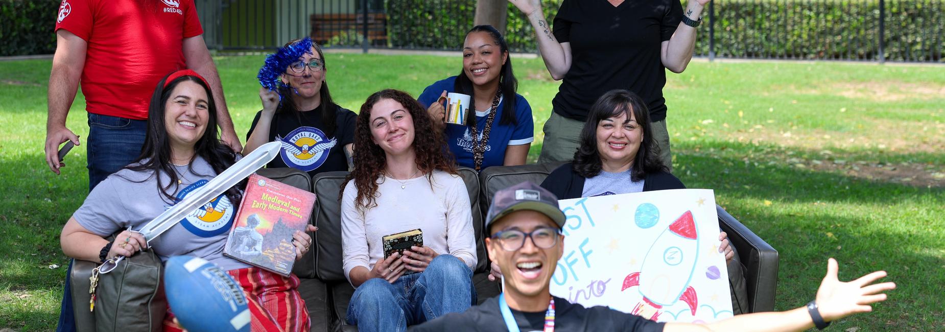 A diverse group of eight adults smiling and celebrating outdoors, with playful props.