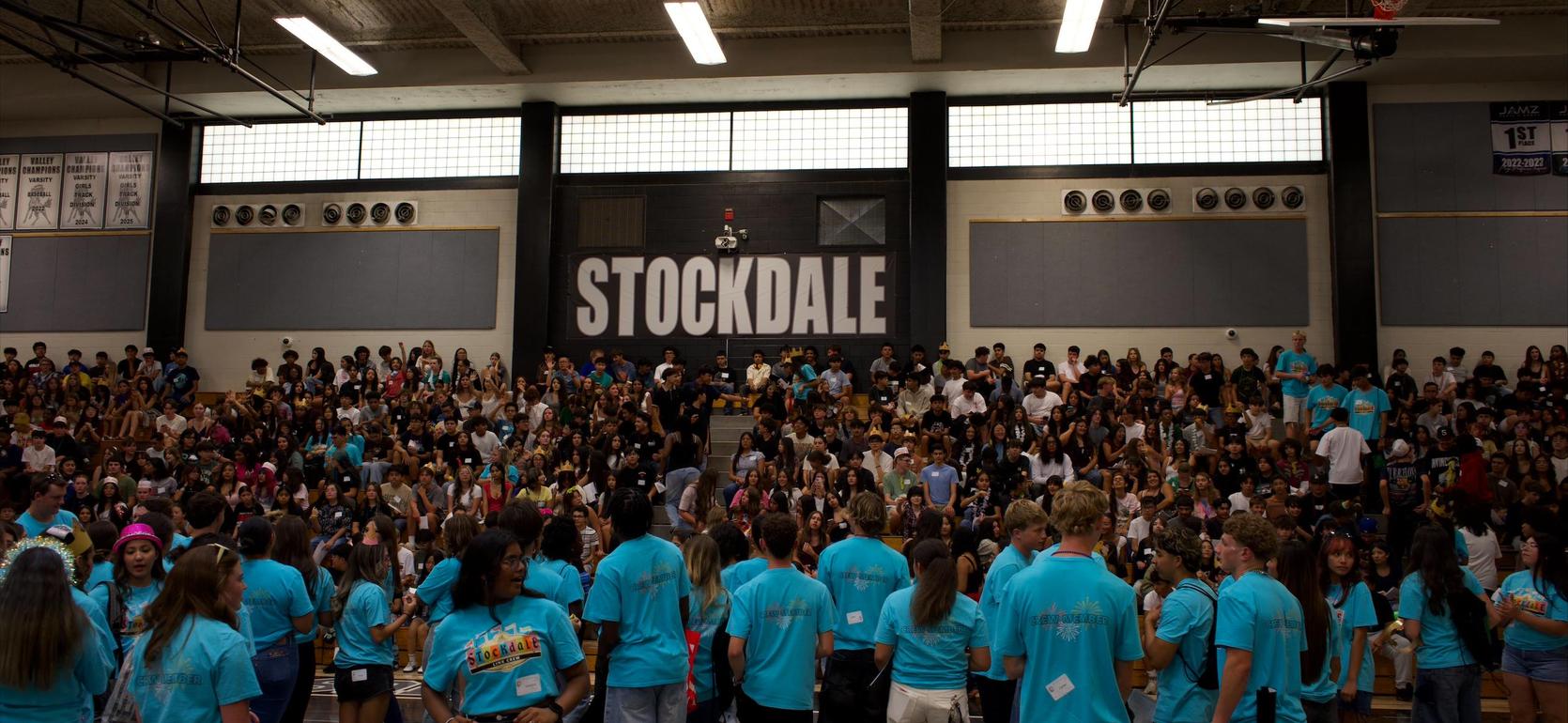 A large group of students in blue shirts gathered in a gymnasium with a 'STOCKDALE' sign.