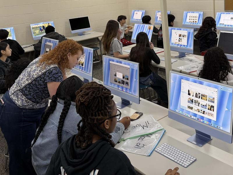 A classroom filled with students at computers, with a teacher assisting them.