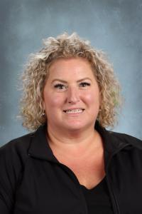 Smiling woman with curly blonde hair wearing a black jacket against a gray background.