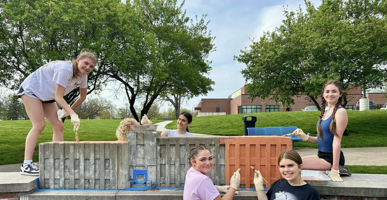Five people painting a stone wall in a park on a sunny day.