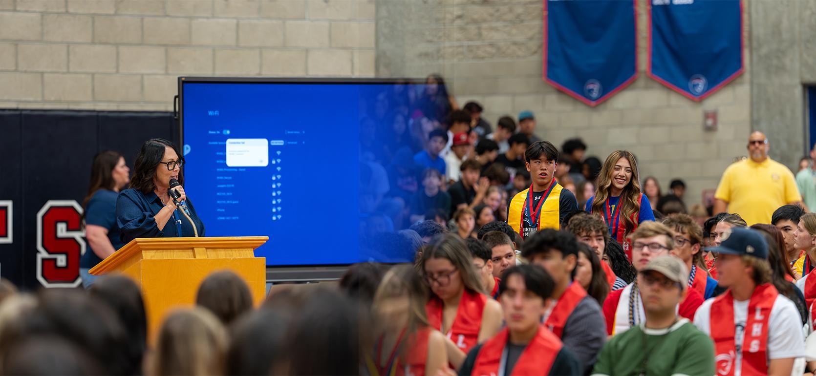 A speaker at a podium addresses a large audience in a gymnasium.