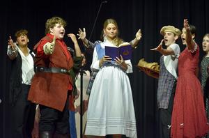A group of young performers on stage singing, with one reading from a book.