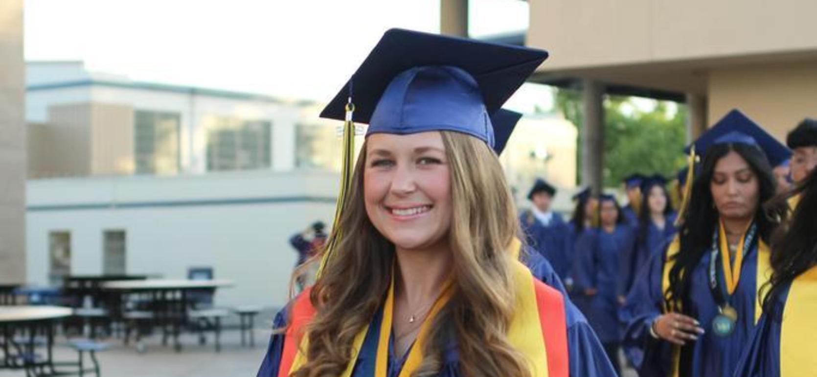 Graduate in cap and gown smiling while walking with peers in the background.