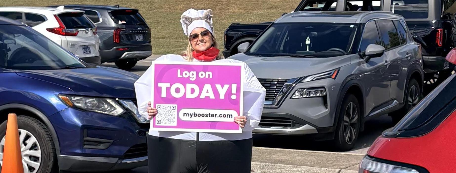 A woman in a chef's hat holding a sign in a parking lot with cars.
