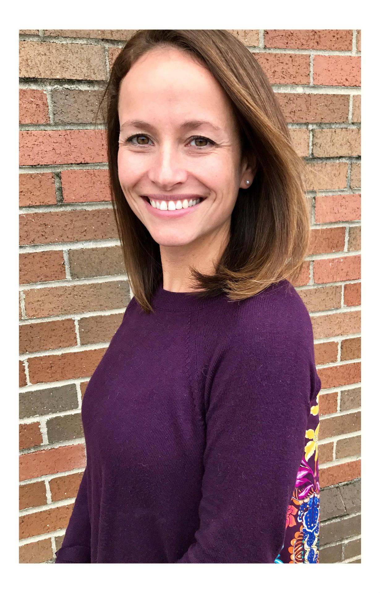 Art teacher, Aimee Price, brown hair and brown shirt in front of a brick wall