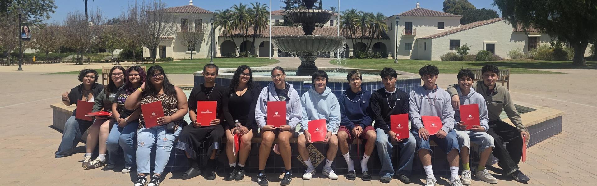 Group of students holding red folders, seated by a fountain and palm trees.