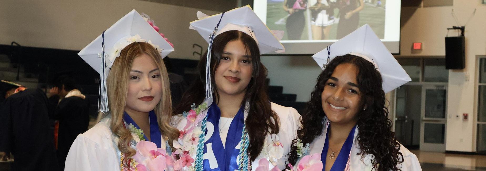 Three female graduates wearing caps and gowns, adorned with leis and sashes.
