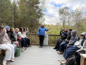 A guide speaks to an attentive group seated on a deck amid trees.