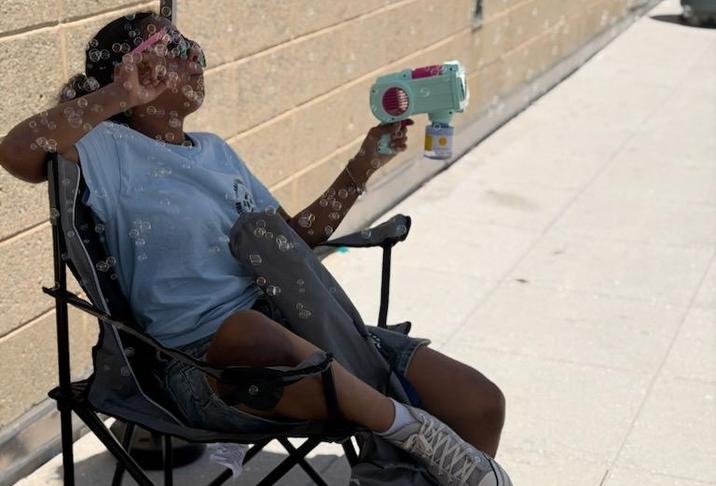 Person sitting in a chair, blowing bubbles with a bubble gun outdoors.