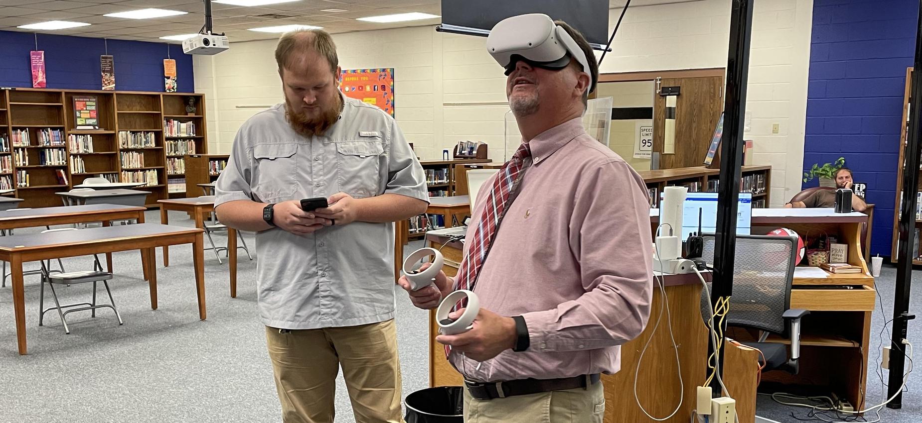 Two men use a virtual reality headset in a library.