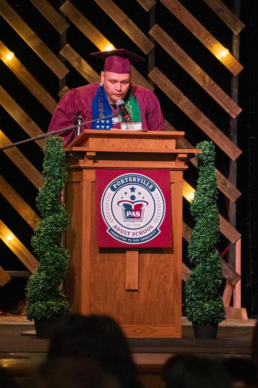 A male graduate in a maroon cap and gown delivers a heartfelt speech from the podium, adorned with colorful graduation stoles and surrounded by celebratory decorations.