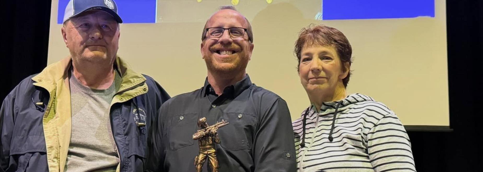 Three individuals smiling, holding an award on stage with a screen behind them.