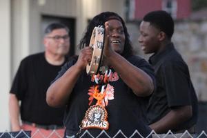 A woman plays a tambourine while smiling, with two men in the background.