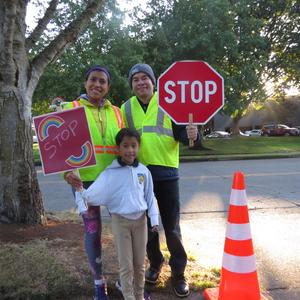 A family at Arco Iris Spanish Immersion School stands outside holding stop signs. They are standing next to a large orange traffic cone.