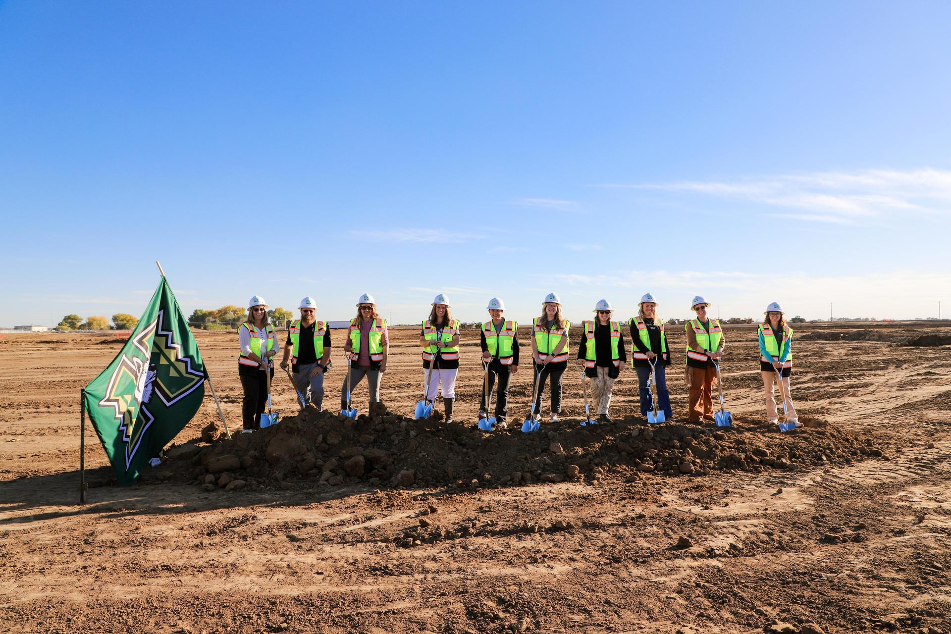 Group of ten staff members with shovels and a flag at a construction site.