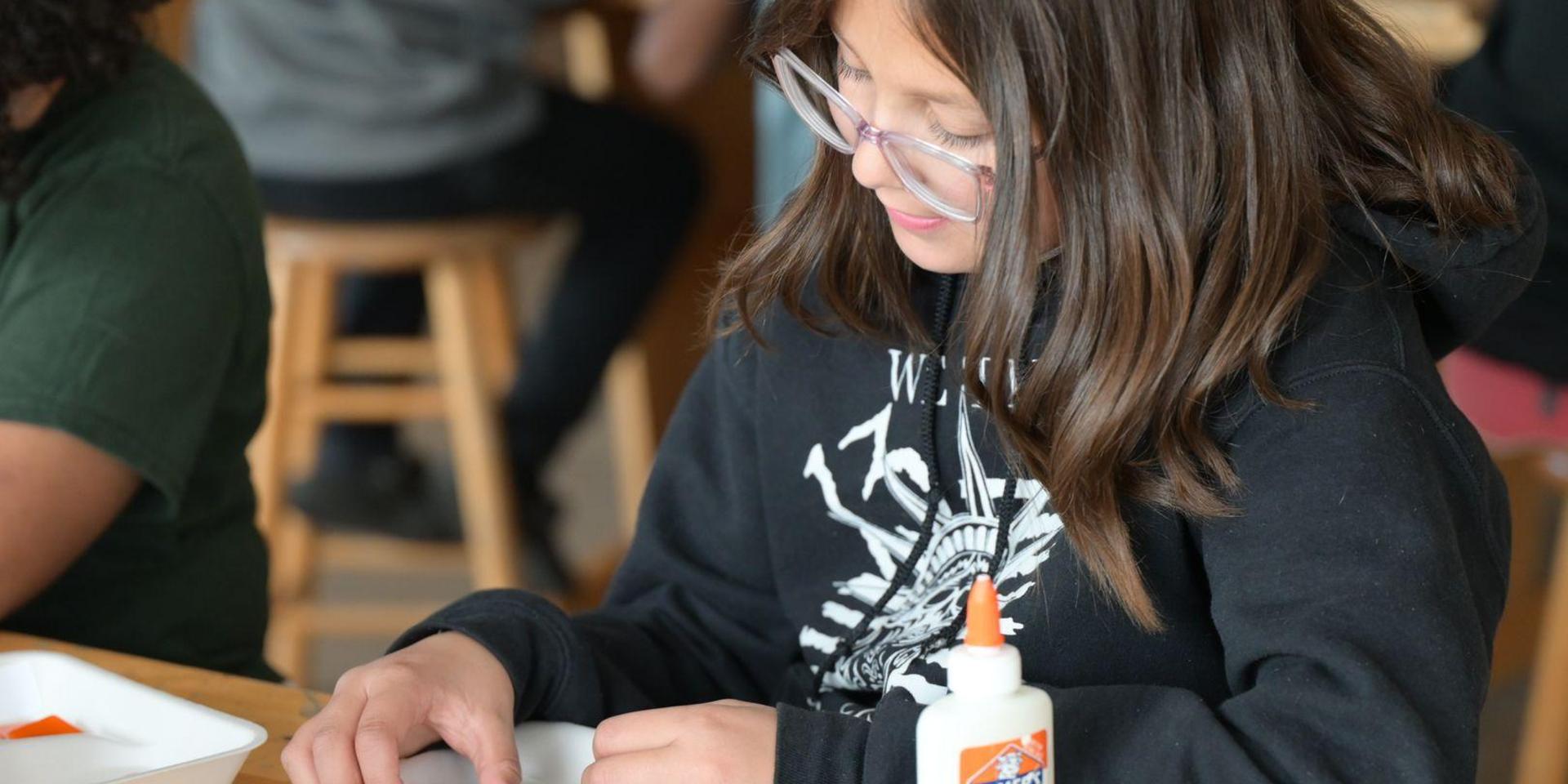 Child with glasses working on a craft project with glue and paper.