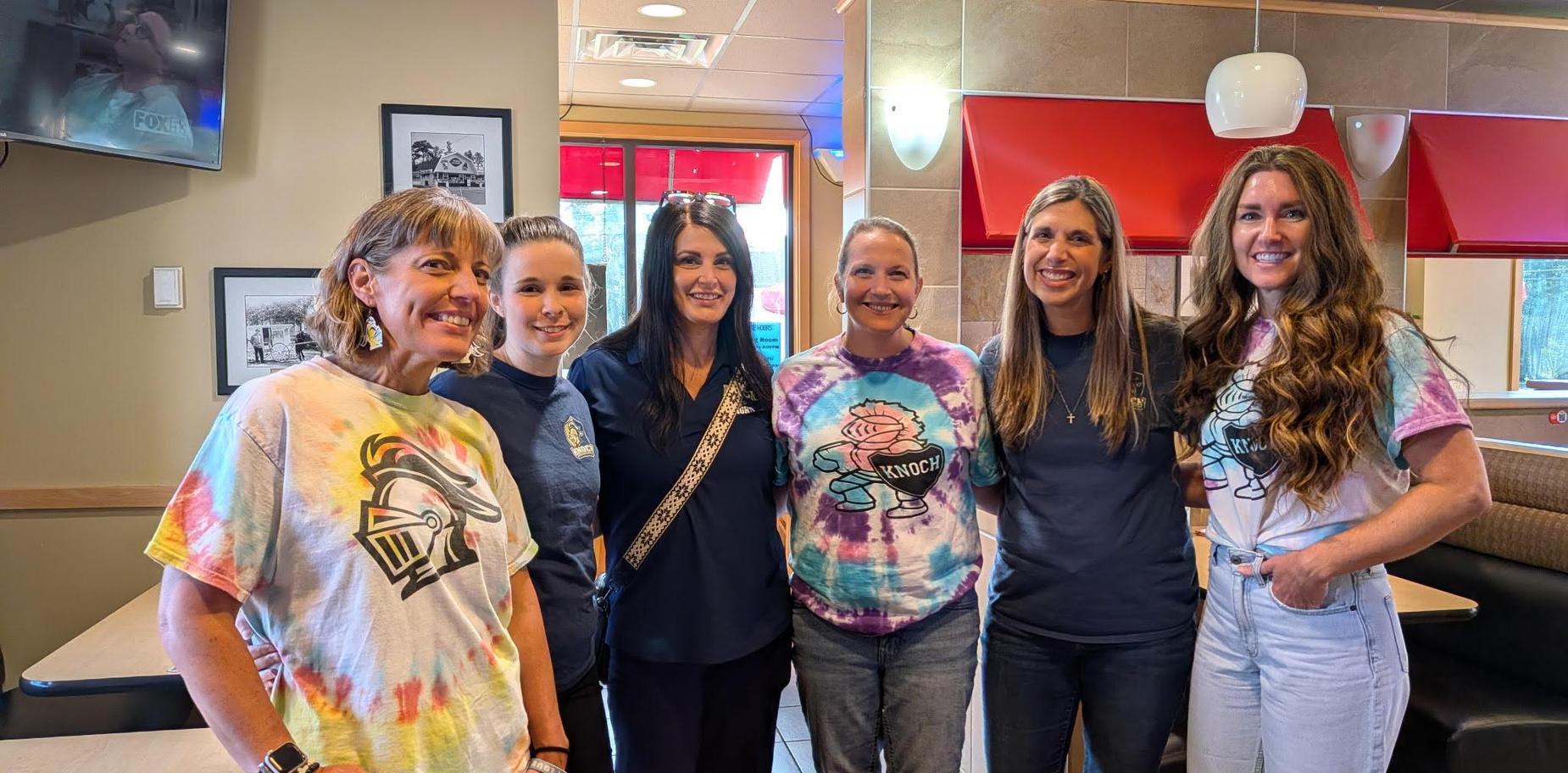 Six women in colorful shirts pose together in a casual dining area.