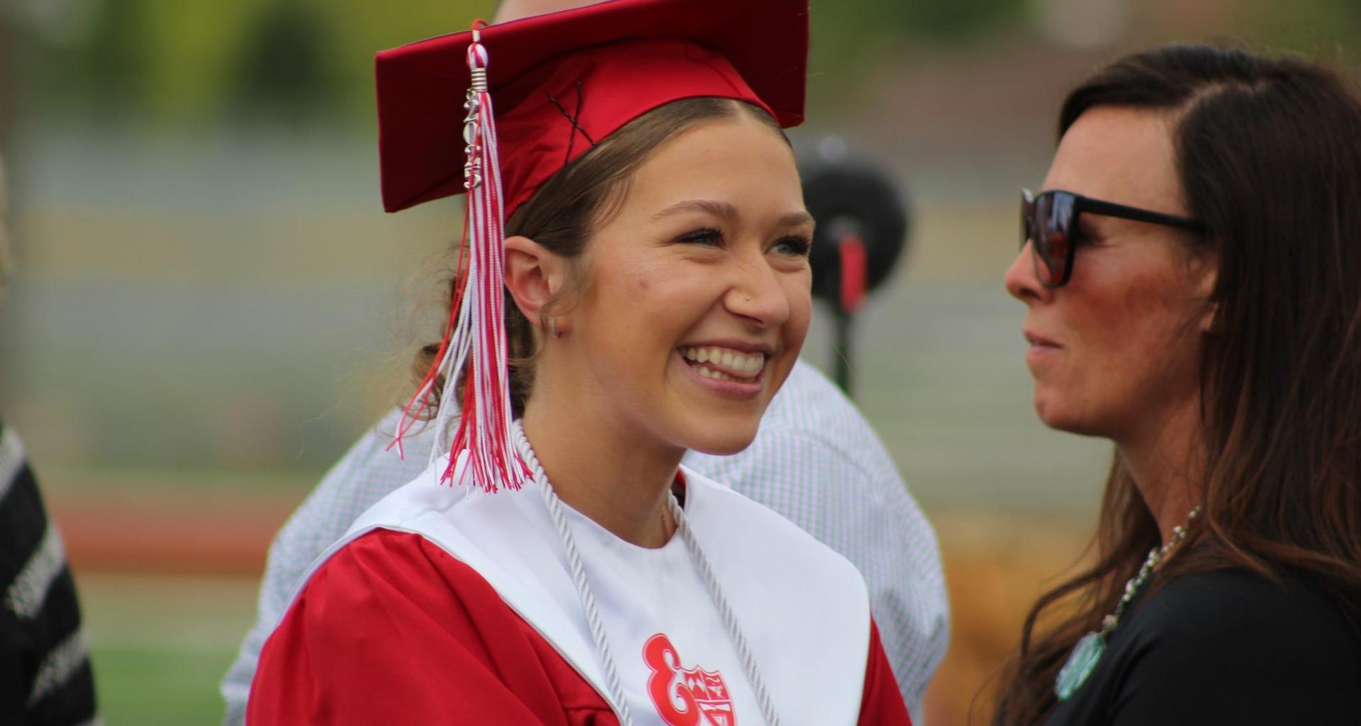 Smiling graduate in a red cap and gown chatting with a woman.