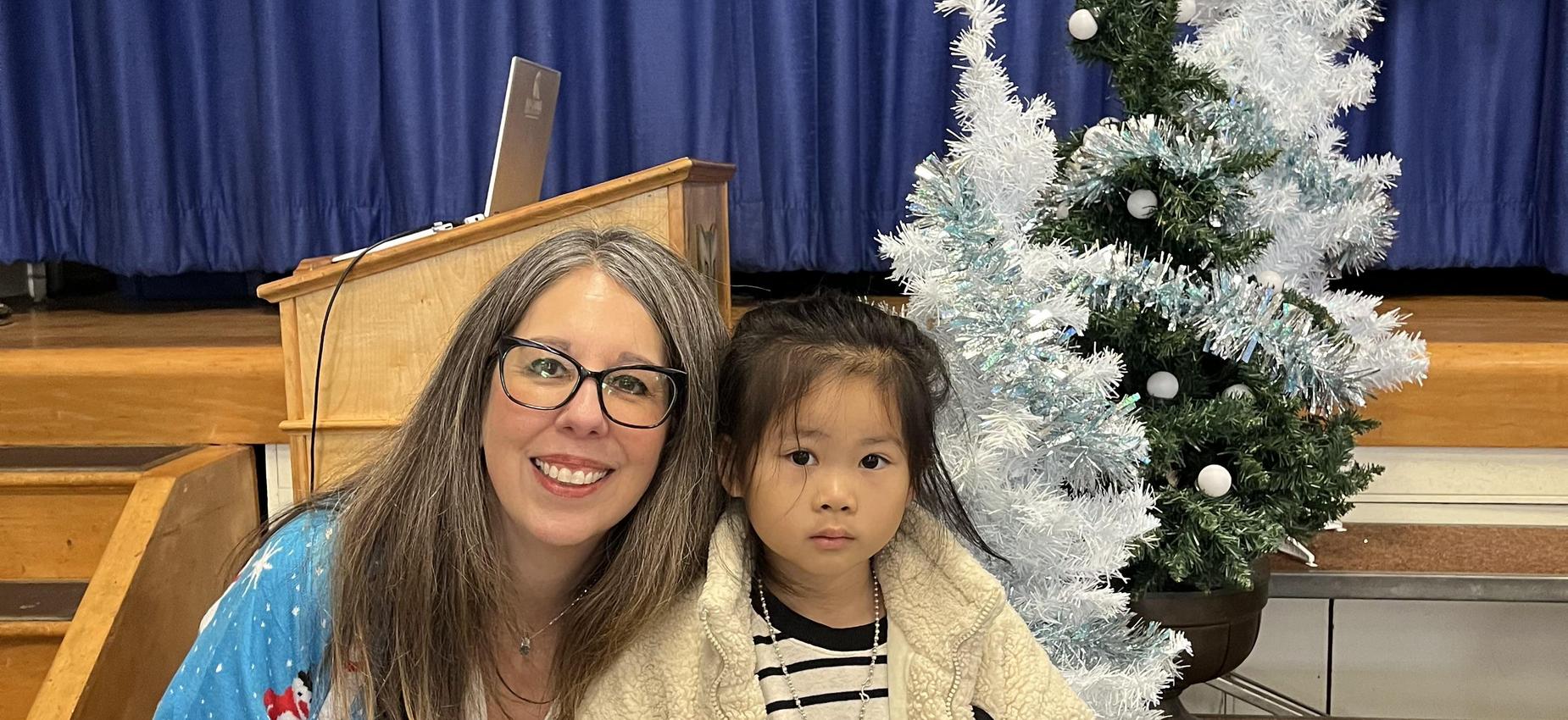 Woman with glasses poses with a young girl beside a white Christmas tree.