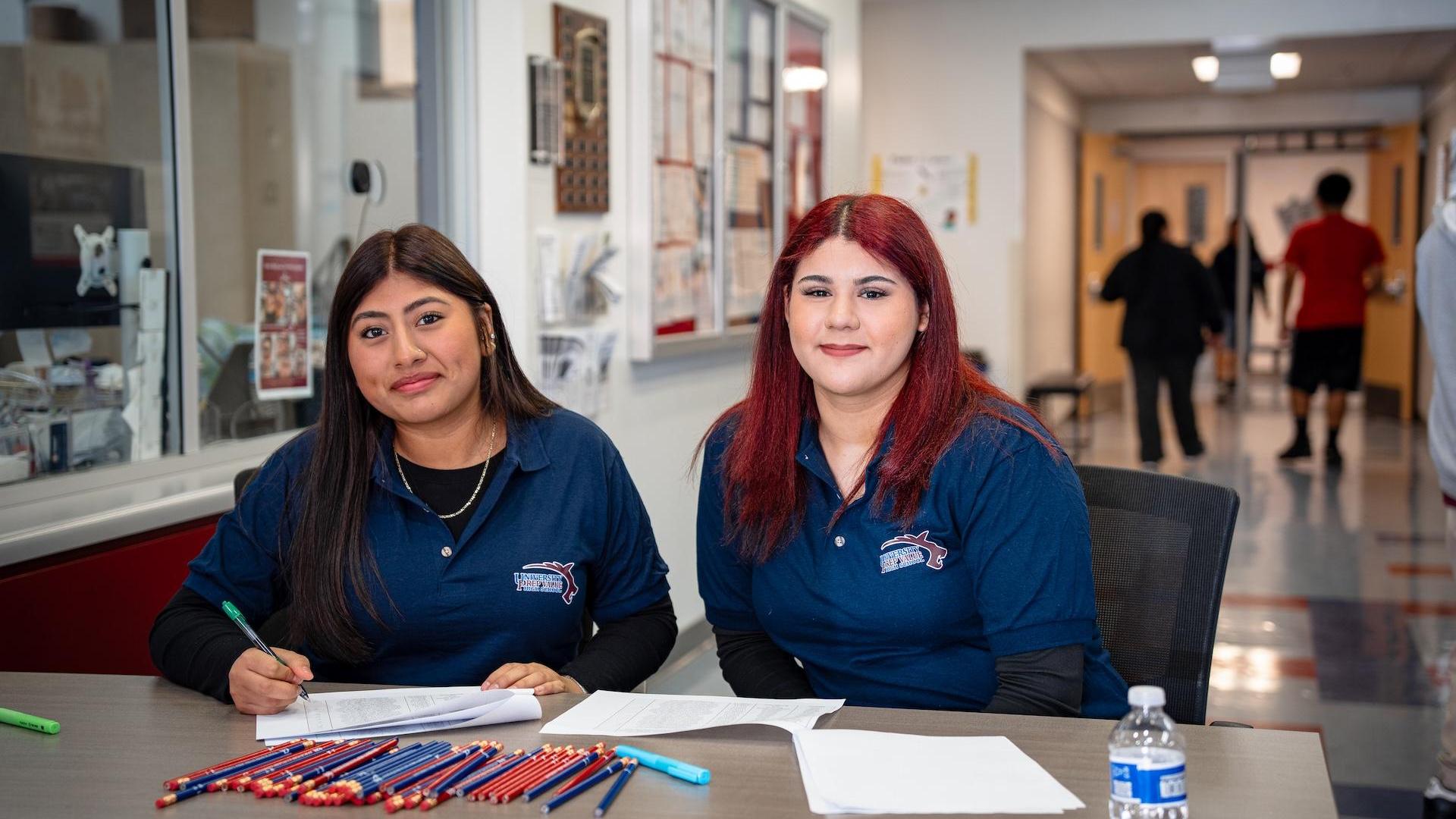 Two women sitting at a table with paperwork and pens in a school office.