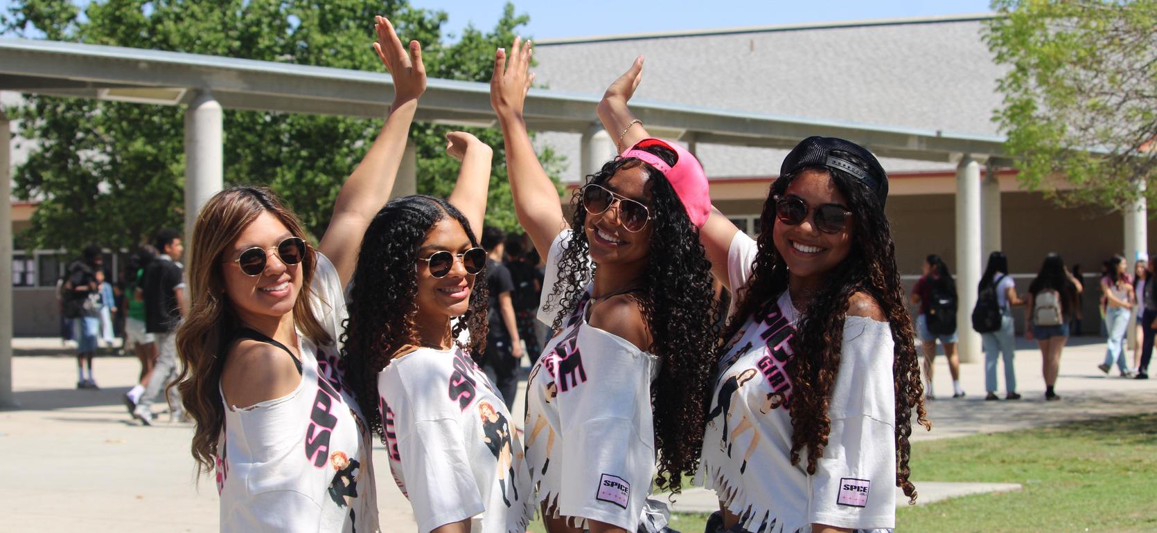 Four young women pose joyfully outdoors with raised hands and sunglasses.