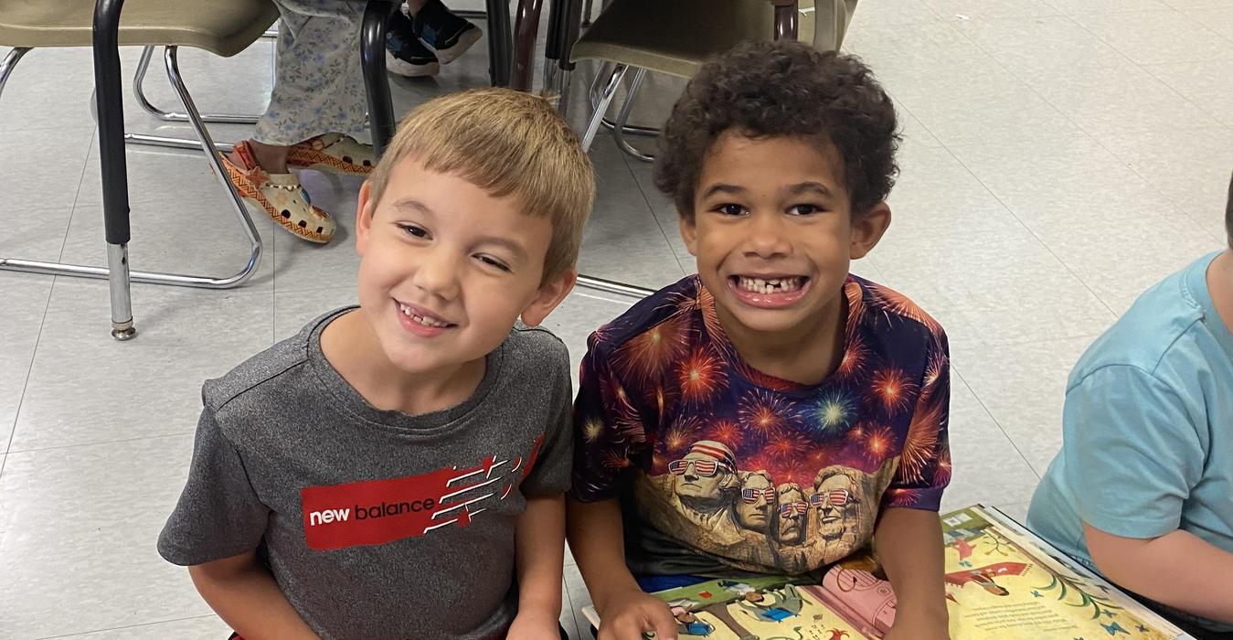 Two smiling boys sitting on the floor in a classroom.