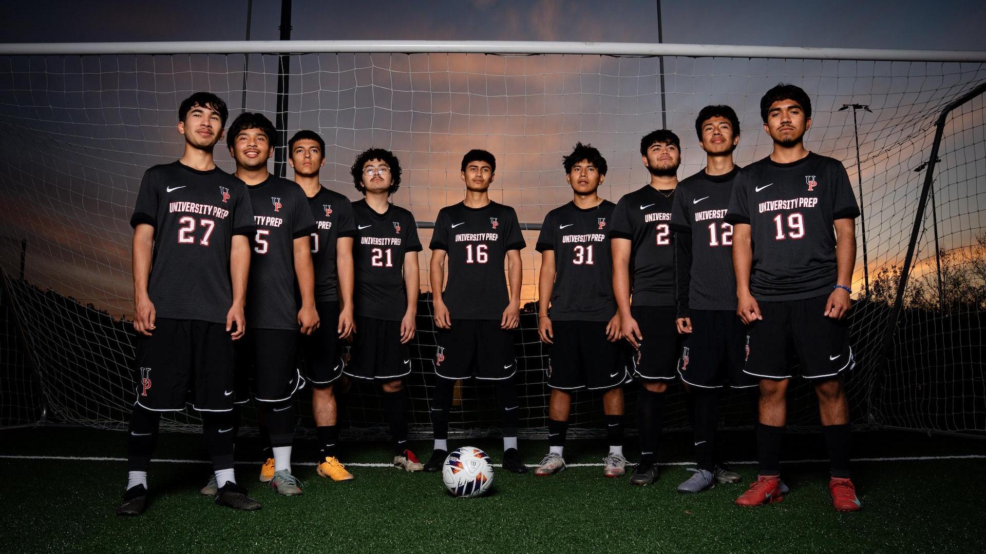 A soccer team in black uniforms poses in front of a goal at sunset.