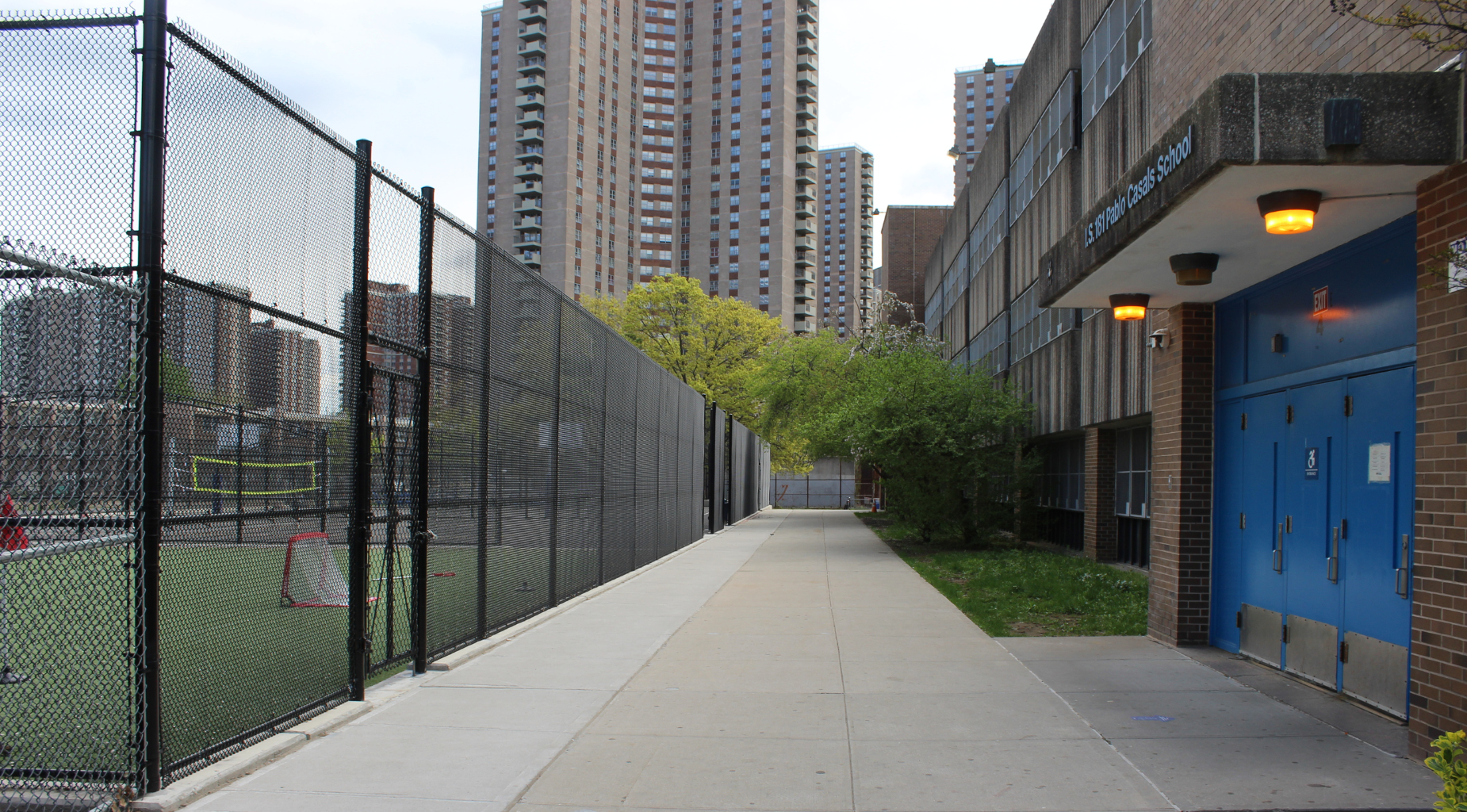 Pathway alongside a fenced sports area and tall buildings.