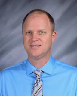 Smiling man in a blue shirt and striped tie against a gray background.