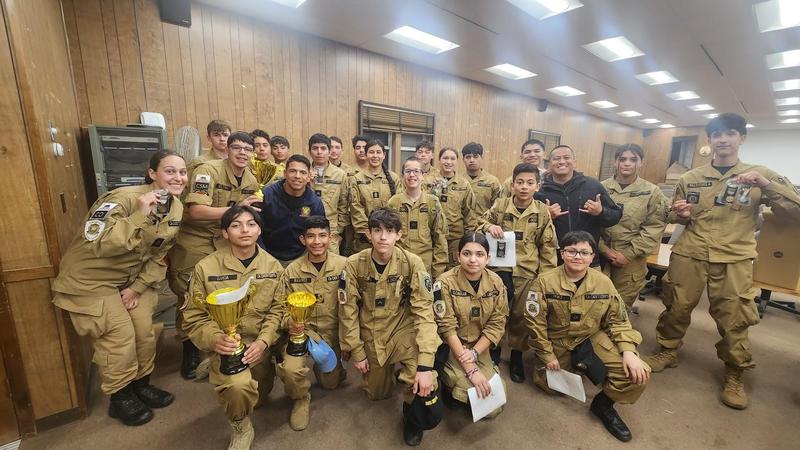 A large group of Porterville Military Academy cadets in uniform pose indoors with big smiles, holding up trophies and medals earned during the marksmanship competition. The students and instructors are gathered closely together, proud of their accomplishments.