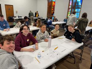 Five QVHS students are sitting around a table where they are working on math problems. All are smiling at the camera.