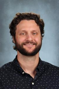 Man with curly hair and a beard, wearing a dark patterned shirt, smiling.