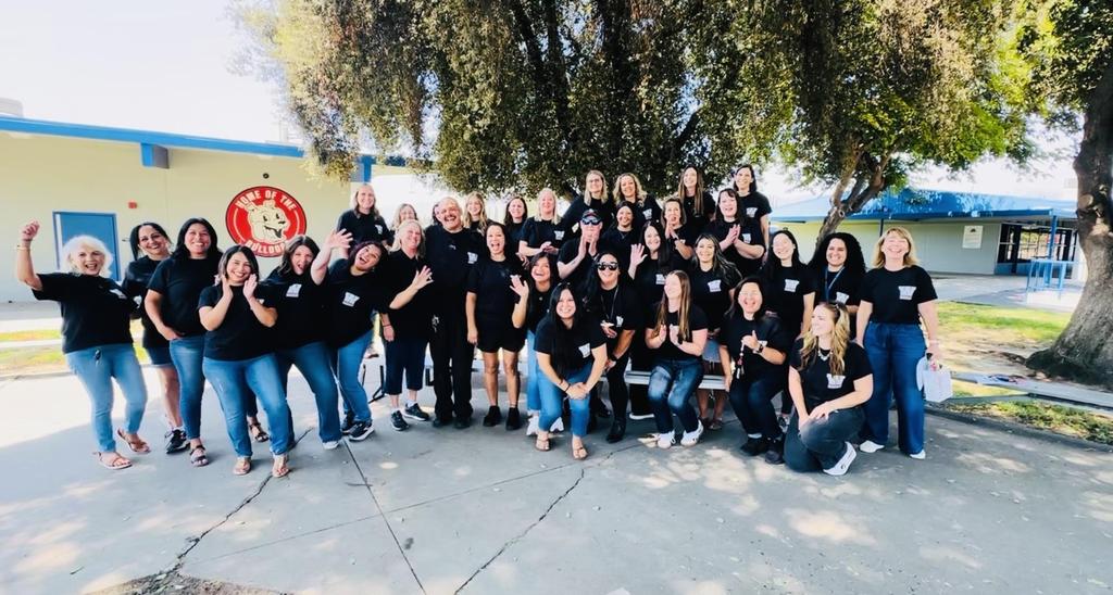 A large group of smiling school staff members in matching black Washington shirts pose outdoors.