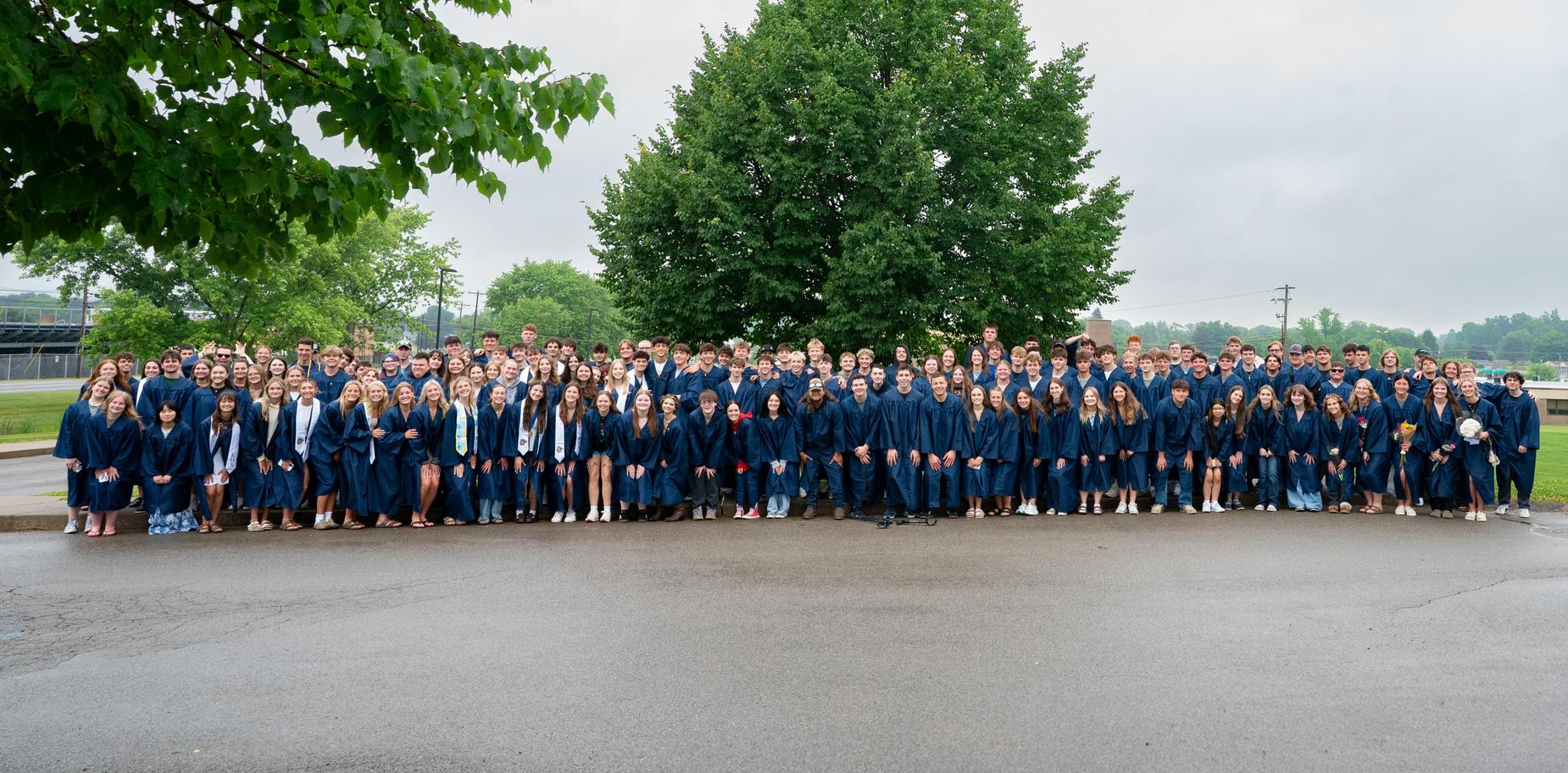 Large group of graduates in blue robes poses outdoors by a tree.
