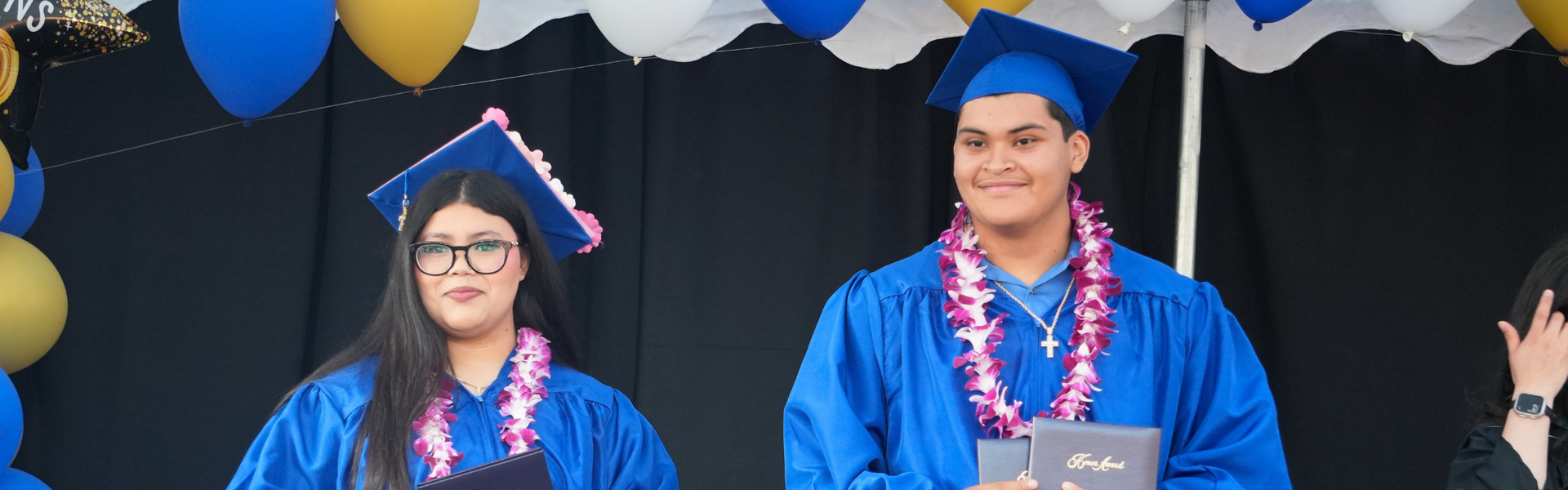 Two graduates in blue gowns with leis, posing for a photo.