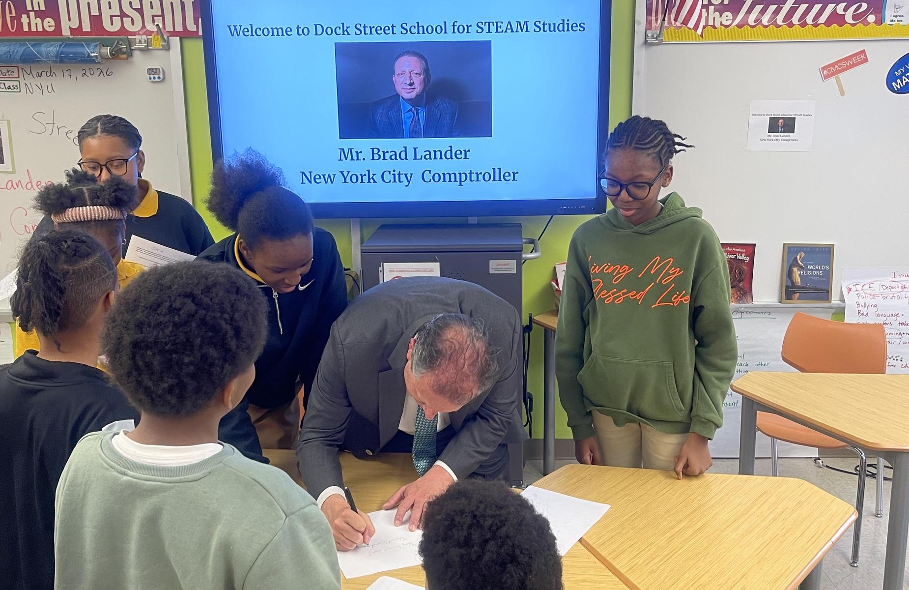 Students gather around a man signing documents at a classroom event.