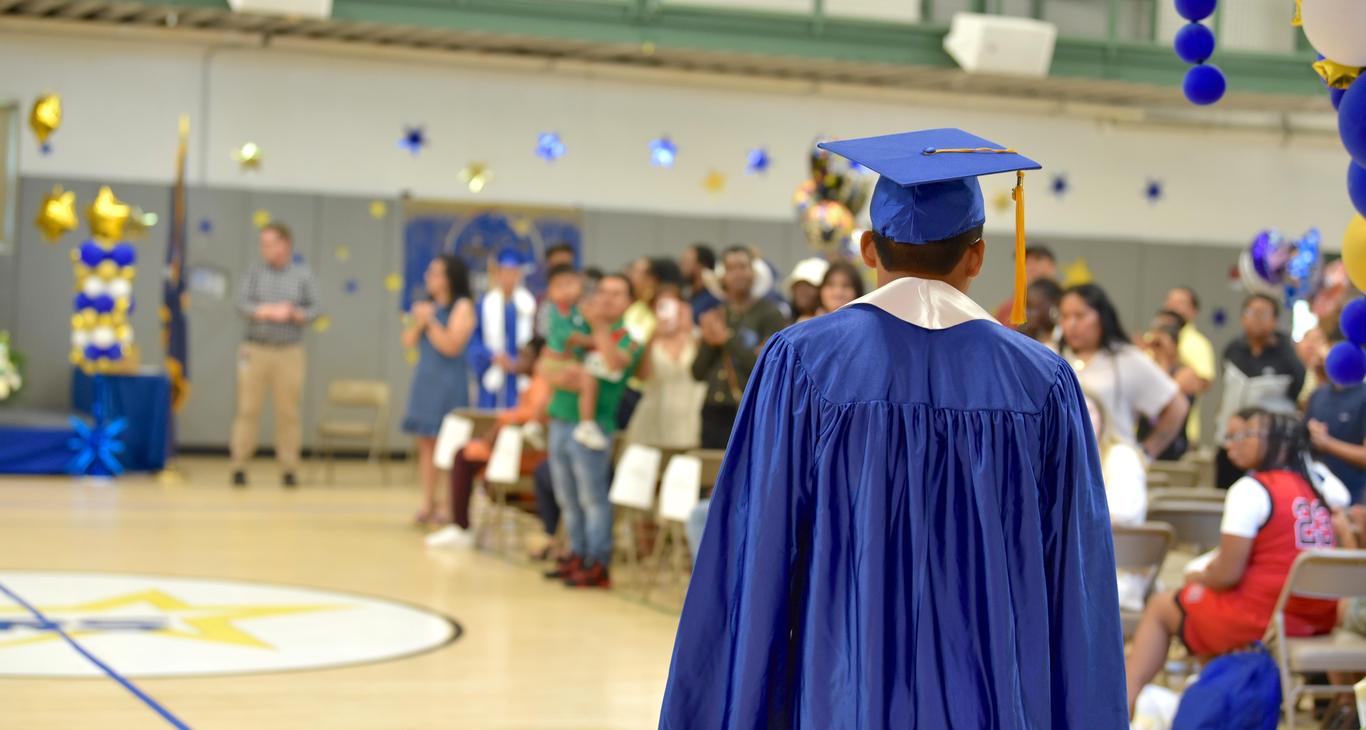 A graduate in a blue gown and cap walks towards a crowd in a decorated gym.